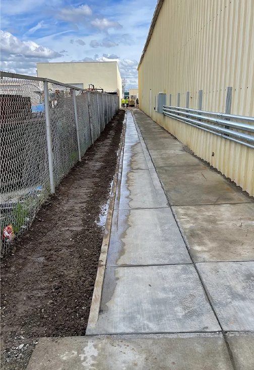 A sidewalk with dirt and grass next to a fence.
